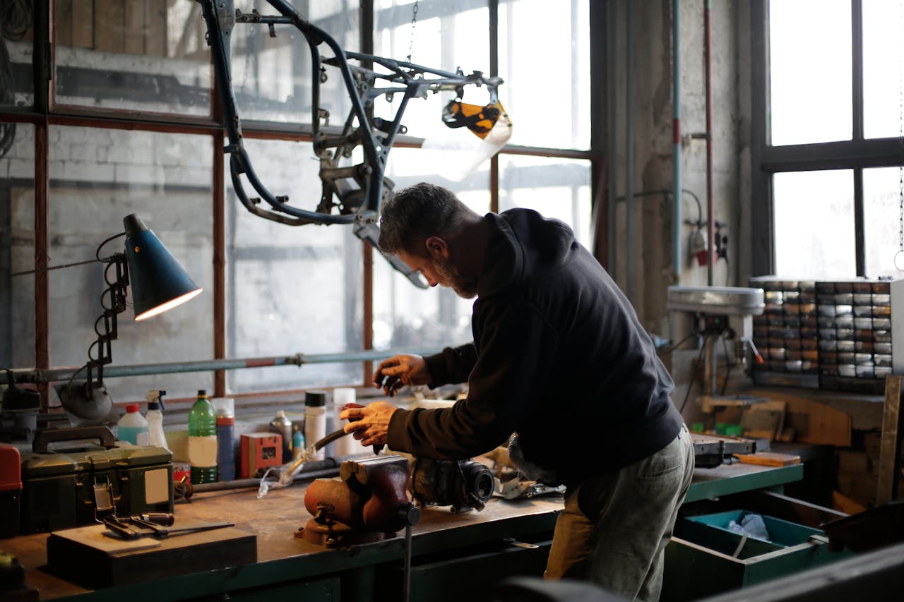 digital-01 Back view of unrecognizable male mechanic in workwear standing near workbench while fixing broken metal detail in lamplight in modern workshop