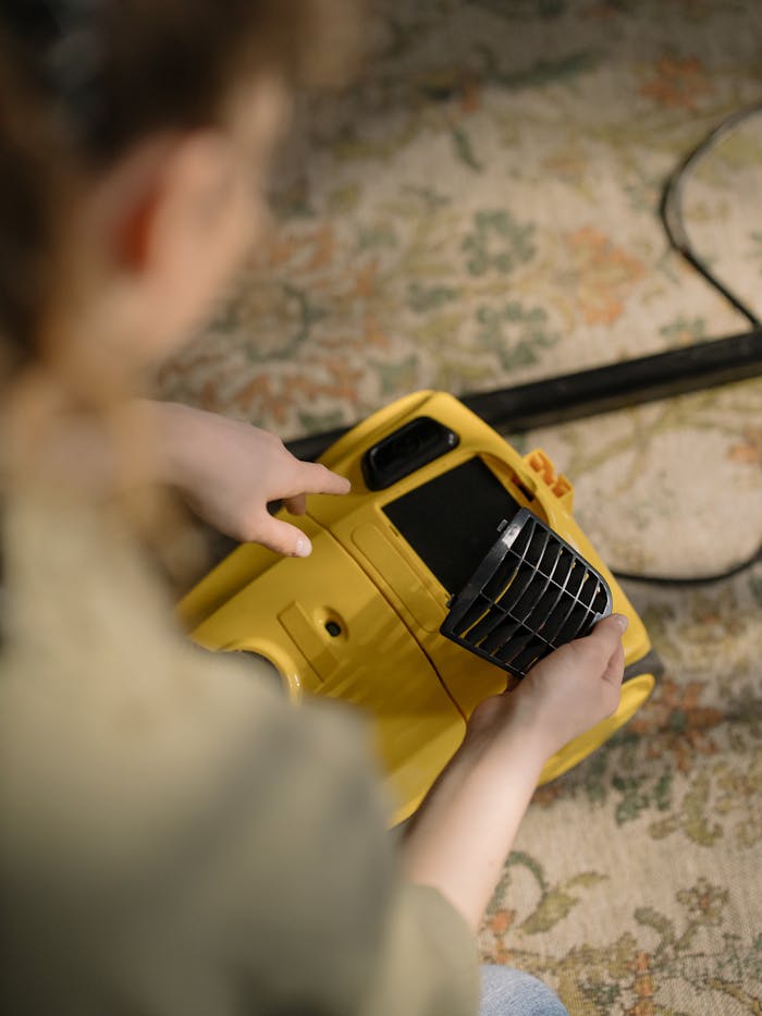brand-03 A person cleans and maintains a yellow vacuum cleaner on a patterned carpet indoors.