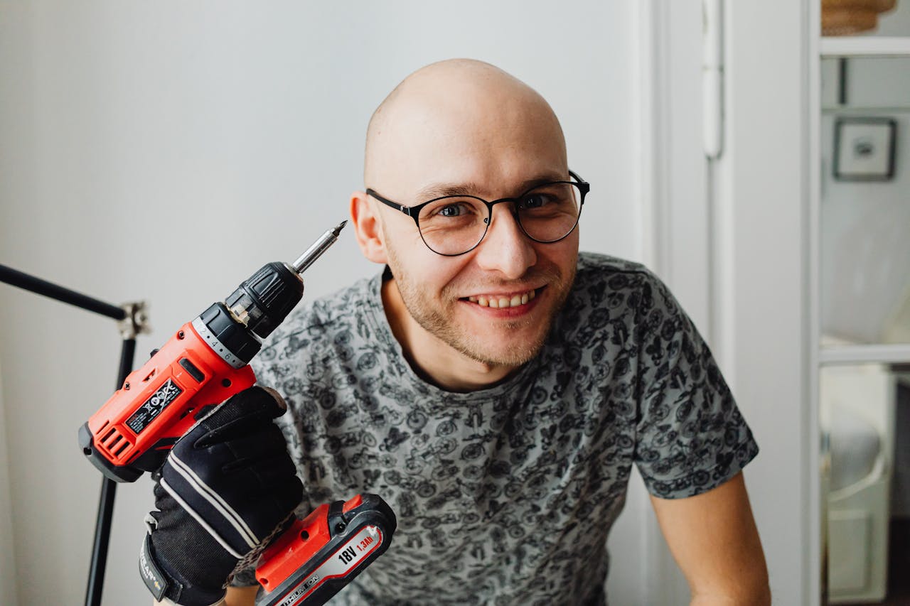 creative-02 A smiling bald man with eyeglasses holding a red power drill indoors.